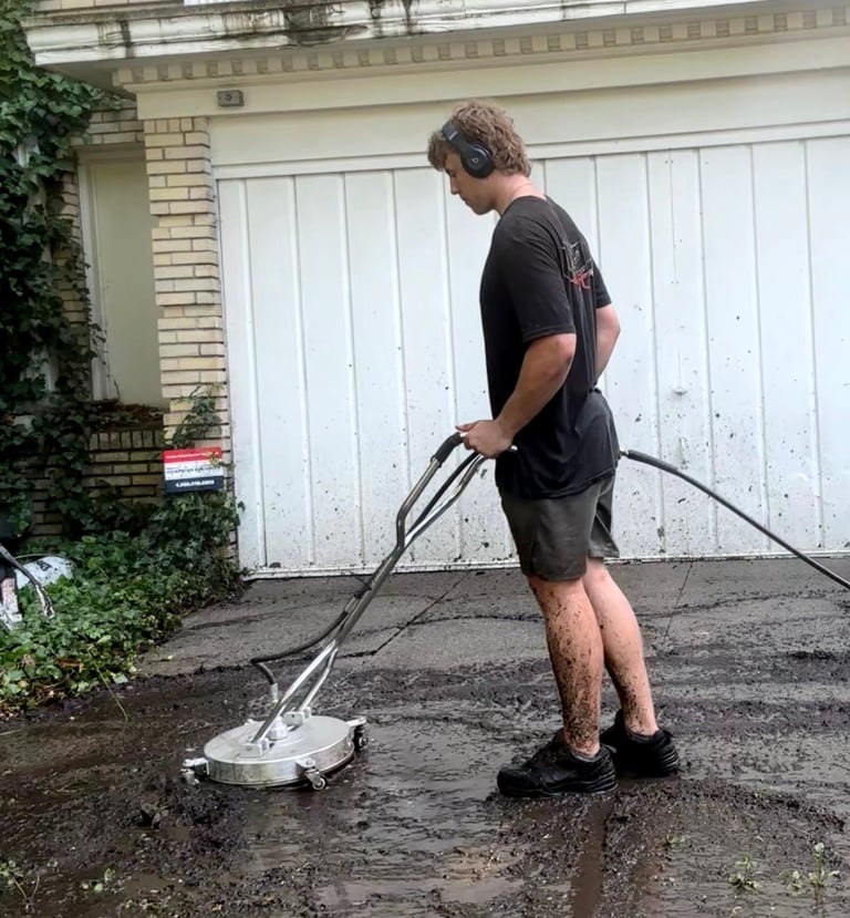 Man using a pressure washer to clean a wet driveway in front of a white garage door