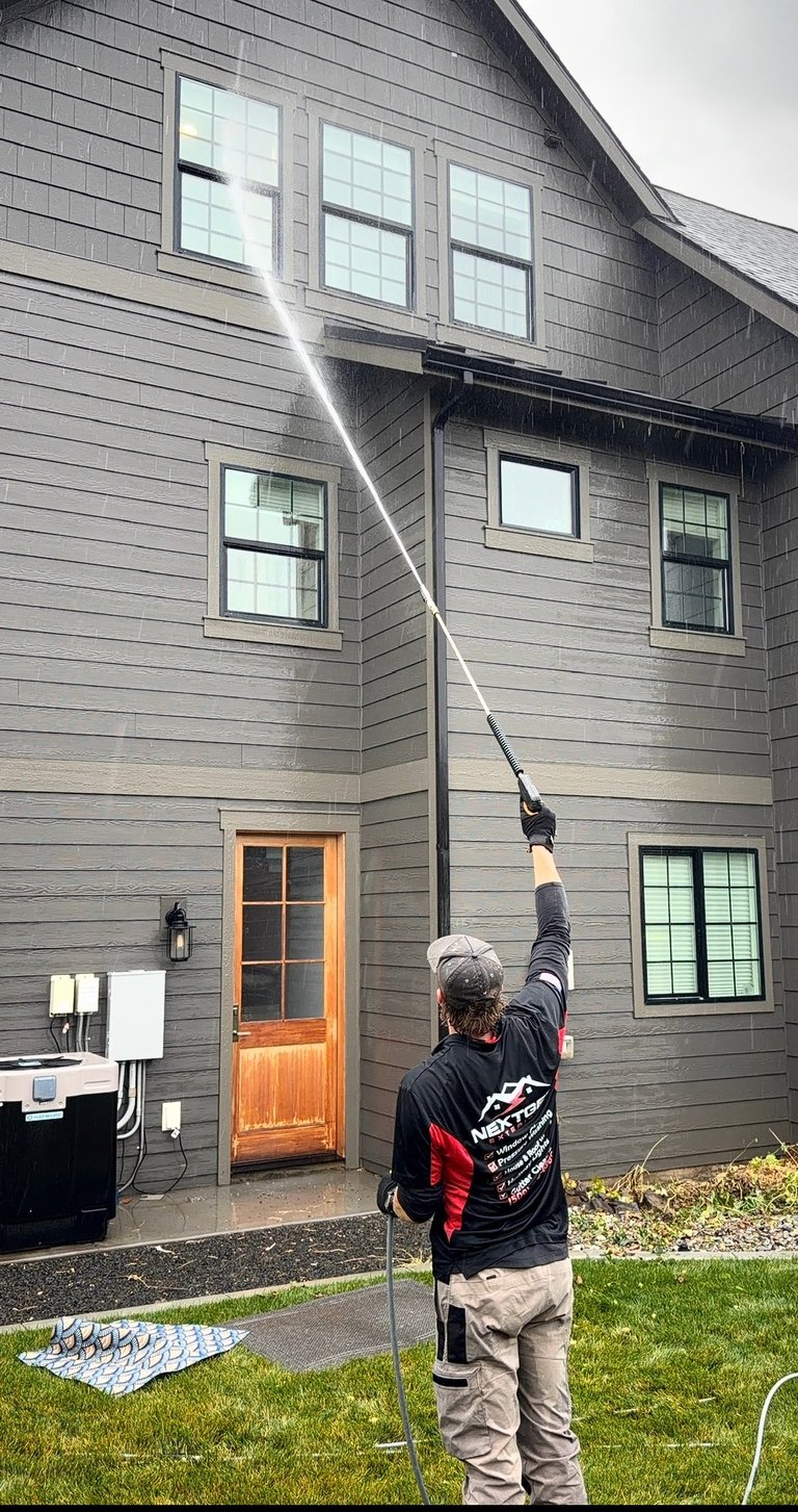 Person power washing the exterior wall of a gray modern house with wooden shingles and multiple windows