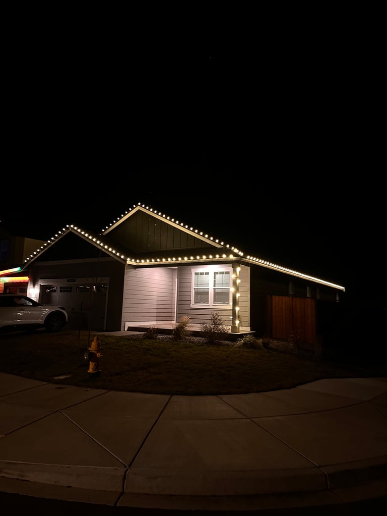 Single-story house with beige siding and attached garage at night, outlined in white holiday lights against a black sky