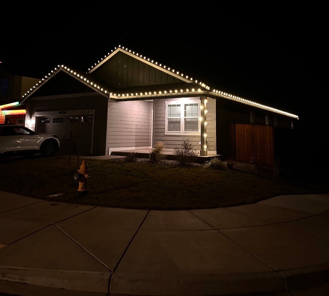 Single-story house at night with warm string lights lining the roof edges, two-car garage on left, front landscaping visible on concrete driveway